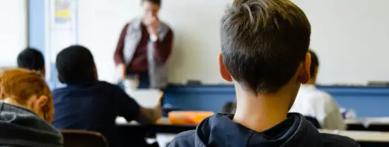  Boy in black hoodie sitting on chair in a classroom looking at the teacher in front