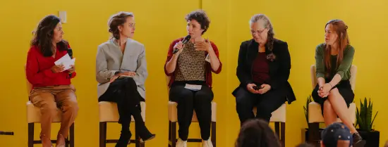 Five women sitting next to each other on high chairs, one is speaking into a microphone